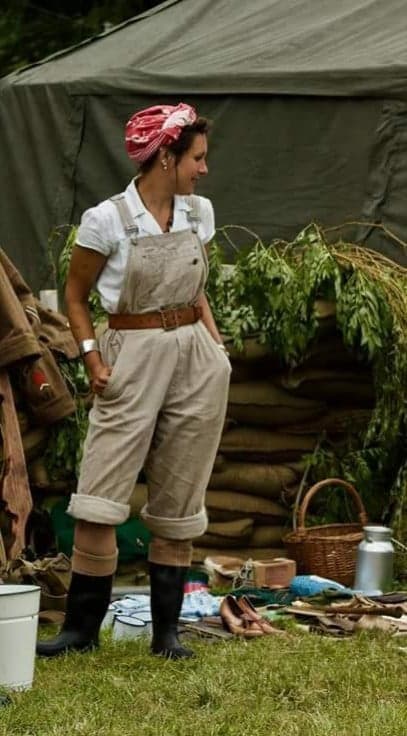 Colour photo of a re-enactment of a WW2 army land girl in beige dungarees and wellies in an outdoor display or gardening and land tools